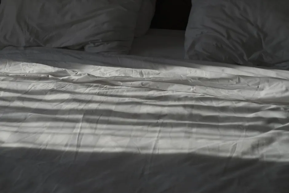 A serene image of white bedding with gentle shadows and two pillows, creating a calm atmosphere.